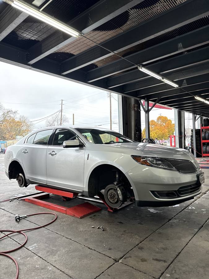 Inside a service bay, a silver sedan raised on a lift with its wheels off, in front of racks of tires and a sign in the background.